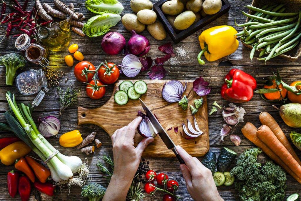 Person chopping an onion while several various vegetables surround the chopping board