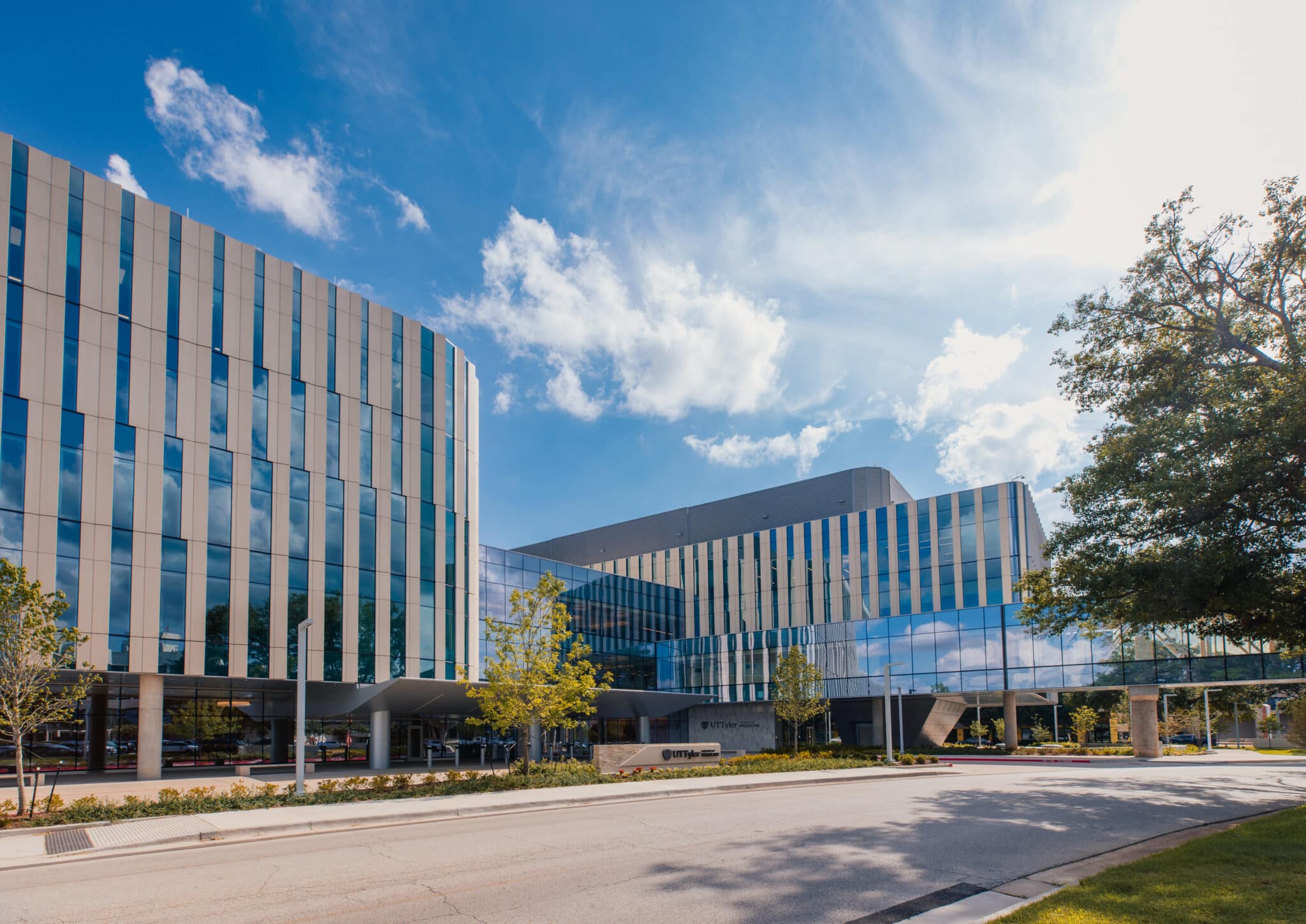 Exterior of UT Tyler medical facility featuring a modern glass facade, landscaped entrance, and blue sky.